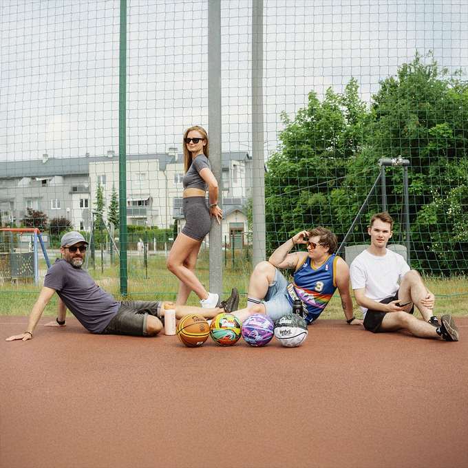 Czwórka znajomych siedzi na boisku do koszykówki w słoneczny dzień, przed nimi leżą różne piłki do gry