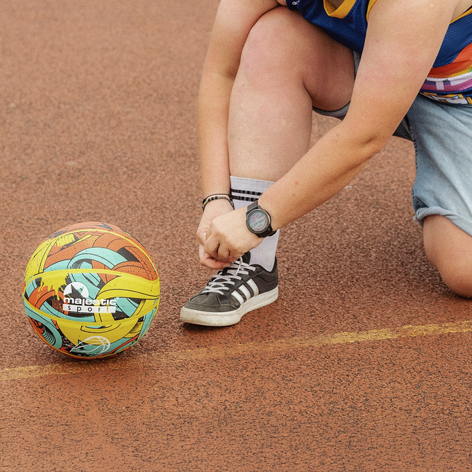 Piłka do koszykówki, Majestic Sport Arrow multicolor, na pomarańczowym boisku do gry obok postaci kobiety